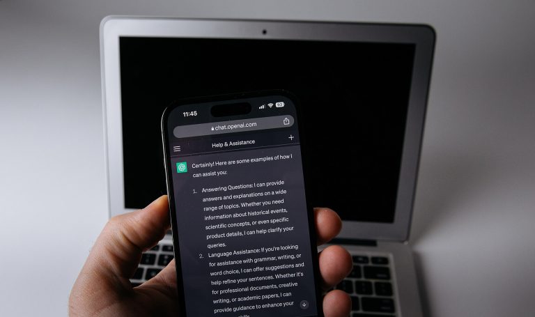 يد تمسك جوالًا مفتوحًا عليه ChatGPT وخلفه لابتوب